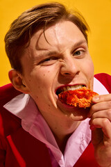 Close-up of young man biting into spicy chicken wing with intense expression against bold yellow background. Concept of fast food aesthetics, pop art photography, emotions
