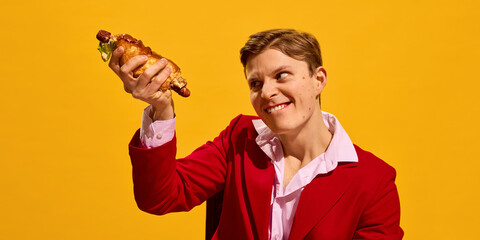 Portrait of emotional young man in red suit with crazy gaze holding hot dog against bold yellow background. Concept of fast food aesthetics, pop art photography, emotions