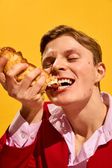 Close-up portrait of handsome young man in red suit passionately eating delicious hot dog against...