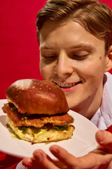 Close-up of handsome young man admiring chicken burger against bold red background. Concept of fast food aesthetics, pop art photography, emotions