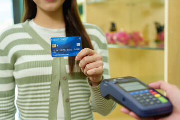 Caucasian adult female customer standing holding blue credit card while Asian fruit shop cashier in denim apron operates contactless payment terminal during checkout at local seasonal market.