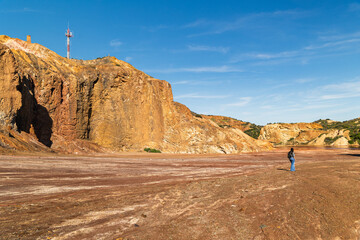 Lone figure in wide oxidized terrain with mineral cliffs and arid vegetation at Mazarrón mines,...