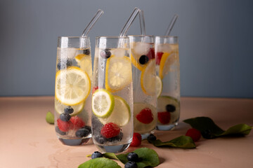 four tall glasses filled with refreshing citrus and berry-infused sparkling water; lemons, limes, raspberries, and blueberries enhance the visual appeal against a minimalistic blue backdrop.