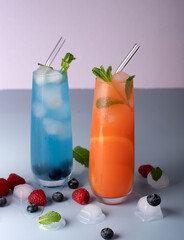 refreshing vibrant summer beverages with blue and orange hues garnished with mint leaves, surrounded by ice cubes and fresh berries on a light background. glassware, clear, simple, crush, tall