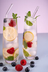 refreshing lemon berry mint infused water in glasses with ice and berries on bright surface against soft pink background, spoon, clear, pastel, white, yellow, red, blue, tasty, delicious, peaceful