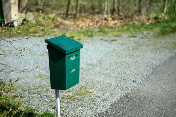 A green mailbox stands prominently on a white post along a gravel driveway. The surrounding area features trees with fresh spring leaves, creating a serene rural atmosphere
