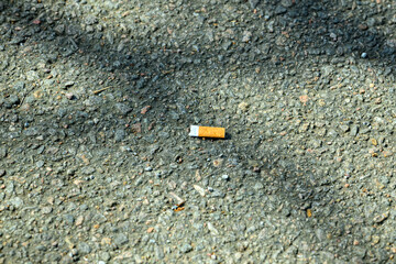 A cigarette butt lies on a gravel pathway, surrounded by small stones and patches of sunlight. This scene captures the impact of littering in urban environments