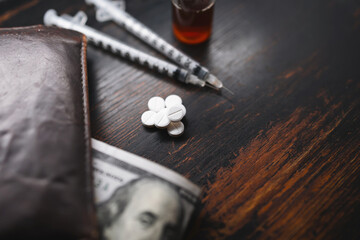 White pills, narcotics or medicine, syringes and glass vial with substance, wallet and US dollar banknotes on a dark wooden table, close-up view