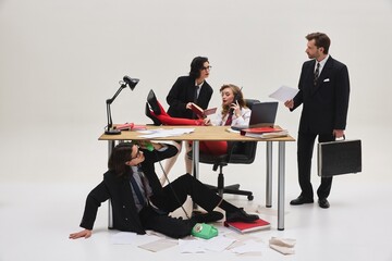 Group of coworkers multitasking with phones and paperwork around chaotic desk in office scene full...