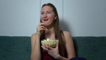 Young woman sitting on green sofa, holding a bowl of popcorn in her hands and watching a sad movie with a melancholic expression on her face and eating popcorn.