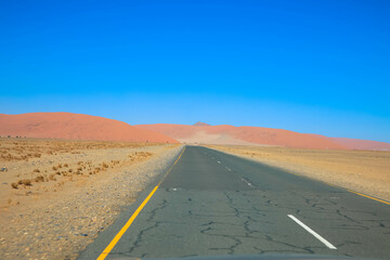 The asphalt road towards Sossusvlei cuts through the rugged and desolate Namibian desert landscape against blue sky. The high red dunes of the Namib Desert rising in the distance. Namibia, Africa.