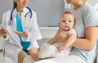 Fototapeta premium Young mother parent sitting with cute toddler child baby and talking with smiling friendly pediatrician family doctor on consultation in clinic and listening recommendations after medical exam.