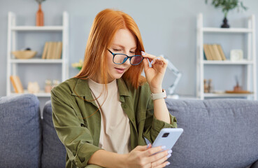 Young redhead woman in eyeglasses sitting on sofa and looking at smartphone screen with surprised...