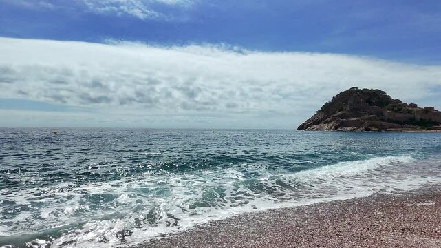 Gentle mediterranean waves washing pebbled shoreline of tossa de mar, spain, under bright blue sky with white clouds and distant small island