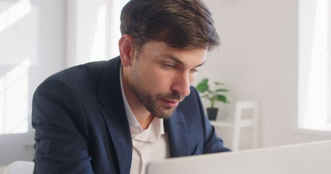 Confident young attractive business man in suit looking seriously at computer monitor screen working at the desk on his workplace in office. Male person at work with laptop. 4k video.