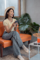 Fototapeta premium Young woman using smartphone sitting on an orange couch wearing a straw hat, smiling and looking away, with a refreshing drink and sunglasses on a side table next to her