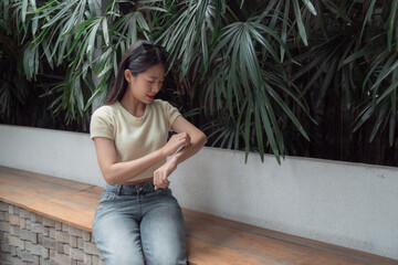 Young woman sitting on a bench scratching her arm suffering from strong allergy outdoors,...