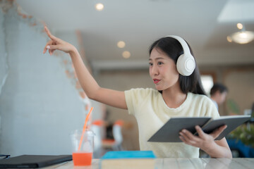 Young student wearing headphones pointing at digital screen during online lesson while holding a book in her hands and sitting at desk in library