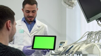 A friendly bearded dentist in a white coat enthusiastically explains treatment options on a tablet to his attentive male patient. The middle-aged Caucasian doctor points to a vibrant green chroma key