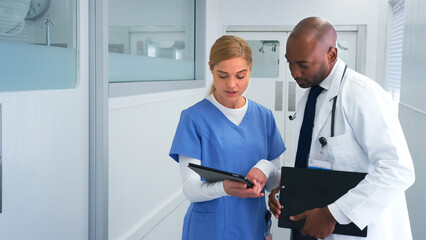 Doctor In White Coat And Nurse In Scrubs Having Discussion Over Digital Tablet In Hospital Corridor