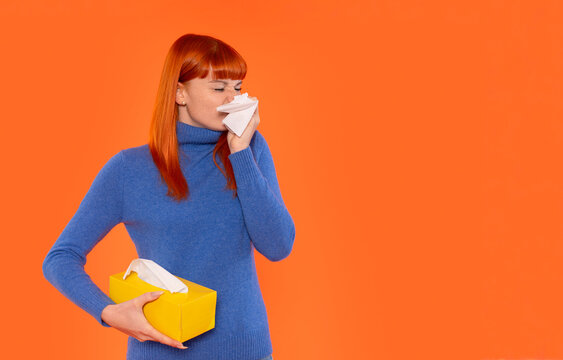 Young woman sneezes while holding box of tissues against bright orange background during allergy season or flu season - Powered by Adobe