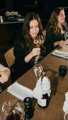 A woman is sitting at a table with a glass of wine in front of her. She is smiling and she is enjoying her drink. There are other people in the background, and a bottle of wine is on the table