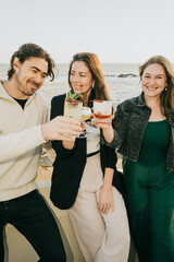 Three people are standing on a beach, smiling and holding up glasses of drinks. The man is wearing a white sweater and the woman is wearing a black jacket
