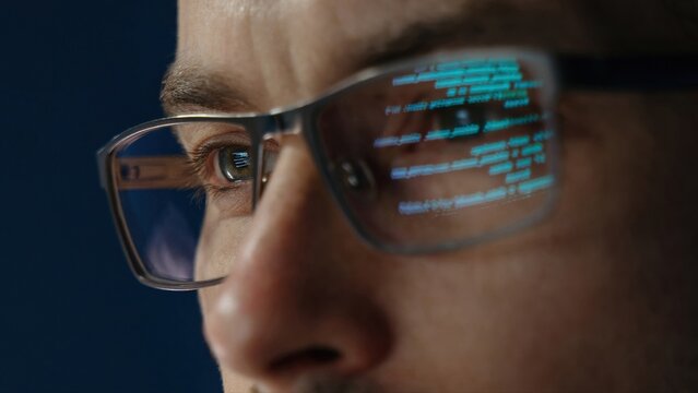 Close up of data scientist face is closely examines data. The glow of code lines reflecting in software engineer glasses and eyes.