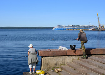 A woman and a man are fishing on the embankment of Lake Onega near the pier with a port crane and a tourist ship