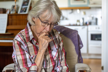 An elderly, gray-haired woman sits, gazing downward with a thoughtful expression.