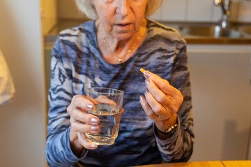 An elderly, gray-haired woman prepares to take her medication, holding a glass of water in one hand...