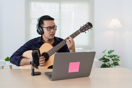Young man in glasses playing acoustic guitar with headphones in home studio environment, recording music on laptop during online session
