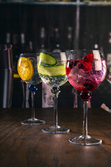 Three stylish cocktails served in large glasses with orange, cucumber and red berries, standing on a wooden surface in a moody bar ambiance, great for concepts of drinks, nightlife or bar aesthetics.
