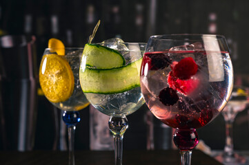 Assorted cocktails with orange, cucumber and berry garnishes served in balloon glasses on a rustic wood surface in a dimly lit bar, ideal for themes like bartending, drinks menu or nightlife.