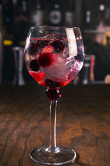 Three elegant gin tonic cocktails with garnishes of orange, cucumber, and red berries on a rustic wooden bar, with a blurry bar background and moody lighting, perfect for mixology or drink concepts.