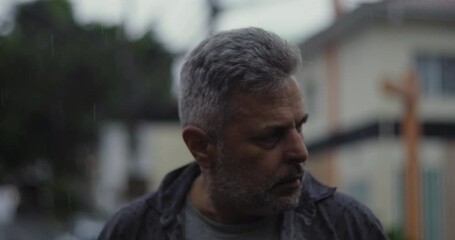 Middle aged man glancing to the side in rain with concerned expression, tense posture and emotional weight in urban street setting - Powered by Adobe