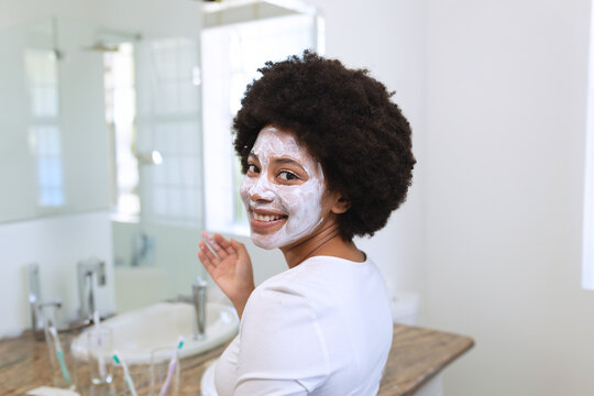 Young African American woman standing at home vanity applying facial mask with mirror, copy space