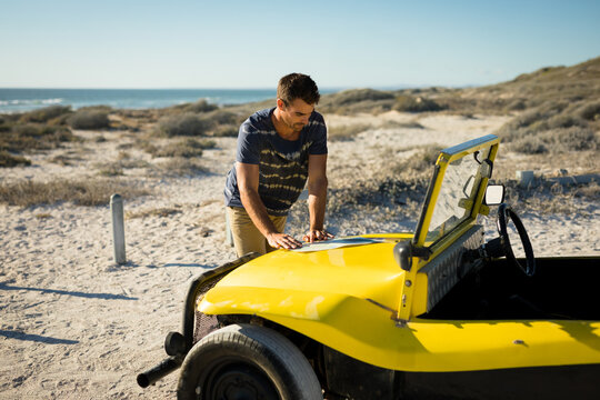 Man leaning and inspecting hood of yellow dune buggy on coastal dune, with ocean view