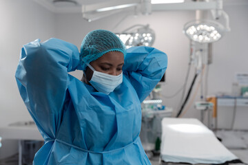 Young African American woman adjusting gown with bouffant cap and mask in operating theatre