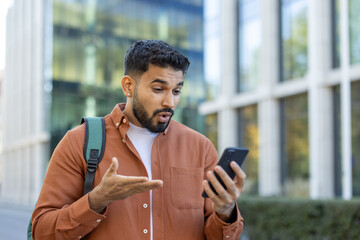 A young man outside reacts with surprise while looking at his smartphone. He gestures with his...