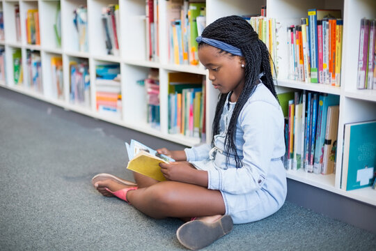 Teenage African American girl sitting on carpet reading small hardcover book in library, copy space