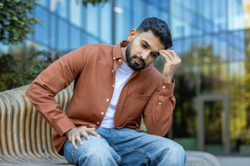 A contemplative man sits on a bench outside, deep in thought with his hand to his forehead. The...
