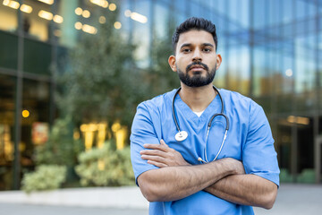 A healthcare professional stands outside, arms crossed, and is wearing scrubs and a stethoscope...