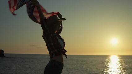 Woman wearing cowboy hat waving american flag, standing barefoot on sandy shoreline during golden sunset, expressing national pride.Usa celebrate 4th july. Independence day concept - Powered by Adobe
