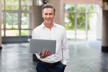 Mature adult man holding laptop in modern office lobby, showing polished floor and greenery