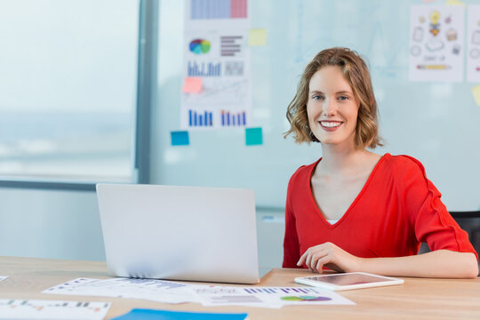 Young woman analyzing data at office desk, with silver laptop white tablet and printed charts