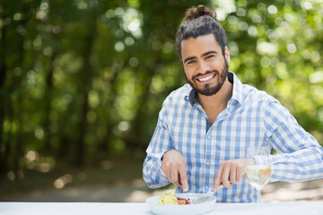 Man sitting at outdoor table in lush forest, cutting salad and sipping wine, copy space