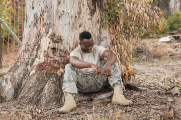 Young African American man sitting against eucalyptus trunk in clearing, wearing military boots