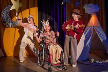 Full length of three young female actors cheerfully singing song along to ukulele on stage, participating in Western musical play at inclusive drama school for children