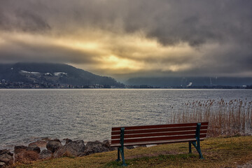 Empty bench on the lake shore against a storming landscape of a lake, mountains, clouds and light